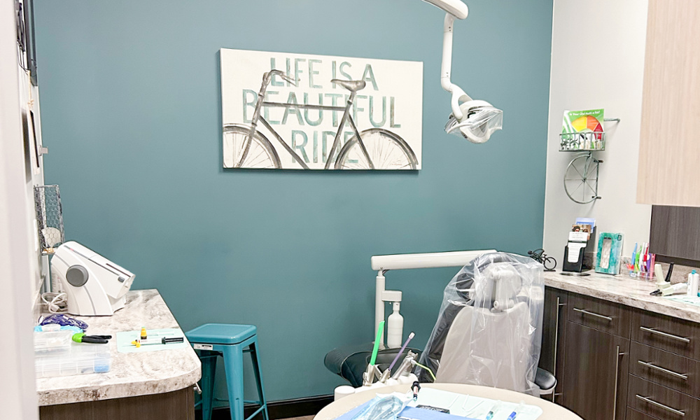 Modern dental treatment room with teal walls, dental chair, overhead light, and bicycle-themed wall art reading 